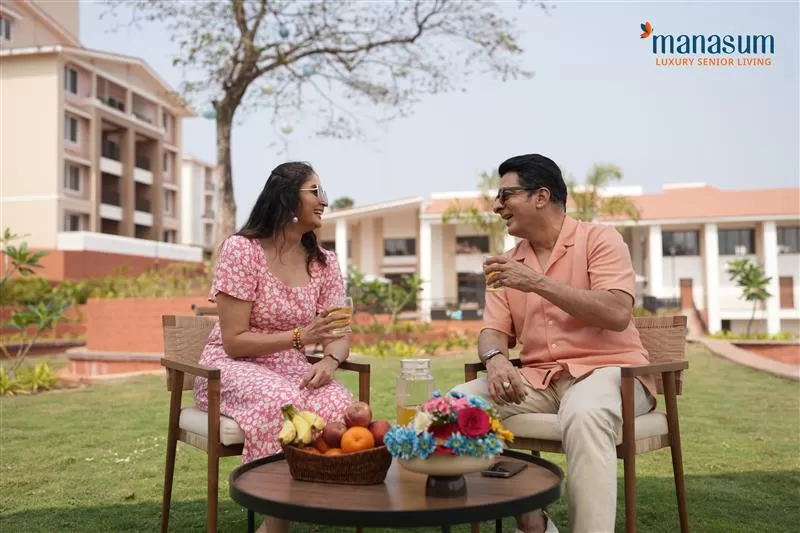 Two adults enjoying refreshments and fruits while sitting outside in a garden area of a senior living community, with modern buildings in the background.