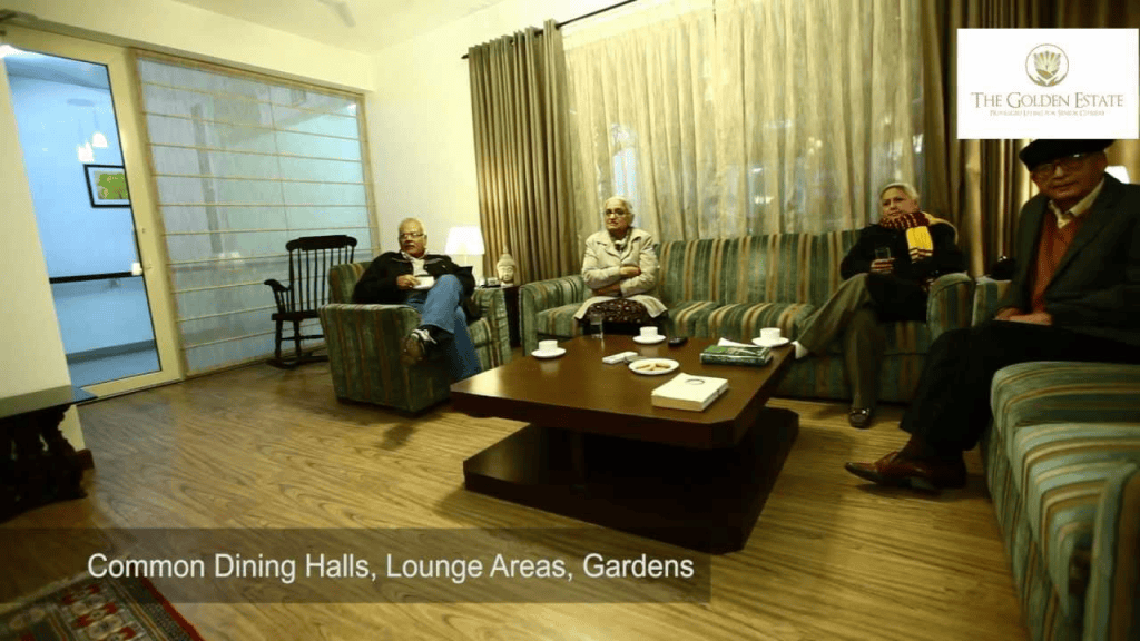 A cozy common area in a senior living community, featuring residents seated comfortably in a lounge with a coffee table, soft lighting, and large windows.