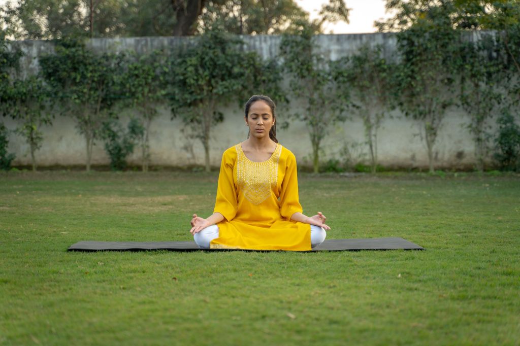 A woman in a yellow traditional outfit practicing meditation on a yoga mat in a grassy area surrounded by greenery.
