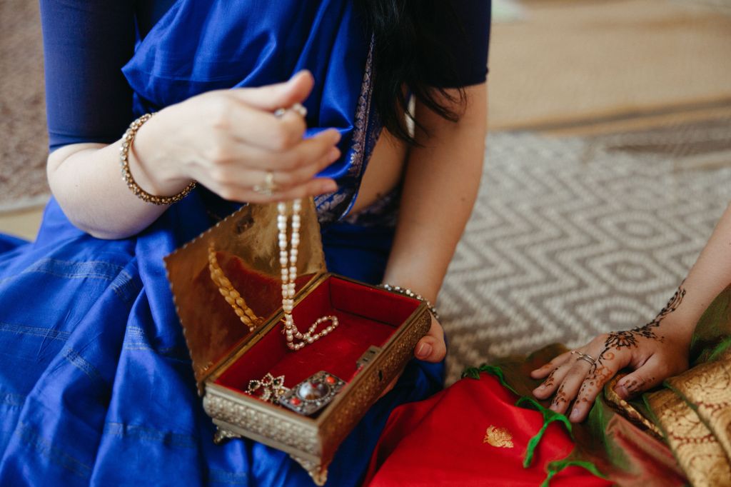 A woman in a blue sari delicately holds a string of pearls while sitting next to a red jewelry box, with intricate henna patterns on her friend's hand visible in the foreground.
