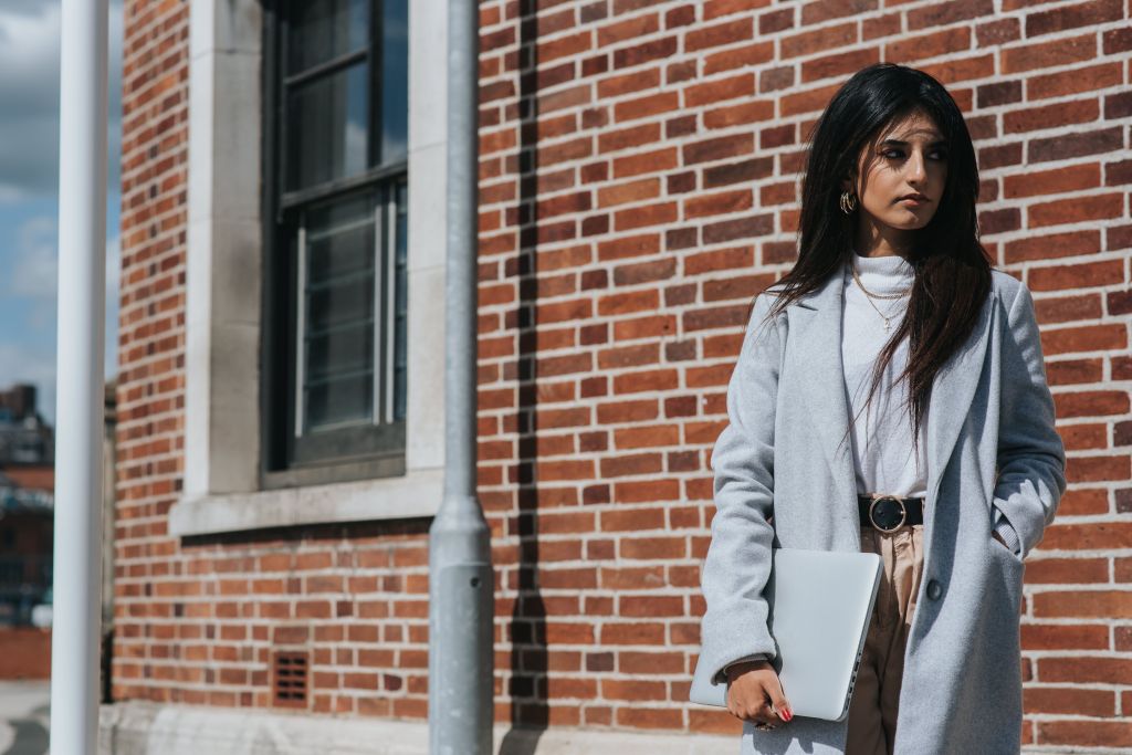 A woman standing against a brick wall, wearing a light gray coat over a white sweater, holding a laptop, with a serious expression.