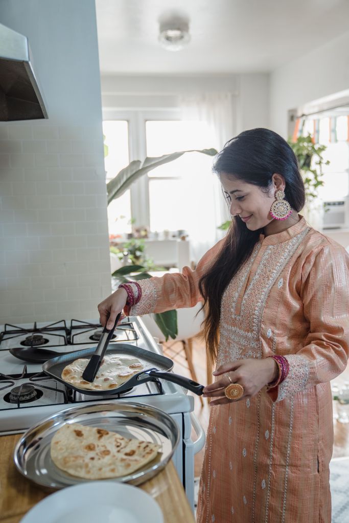 A woman in traditional attire cooks flatbreads on a stovetop, using a spatula. The kitchen is bright and homey, with plants and natural light.