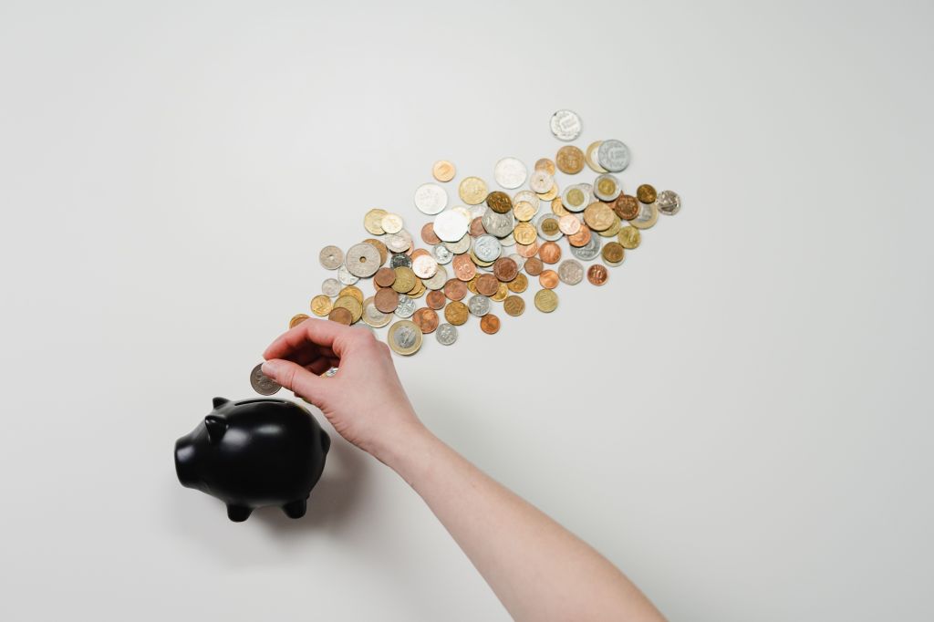 A hand placing a coin into a black piggy bank, surrounded by a scattered collection of various coins on a white background.