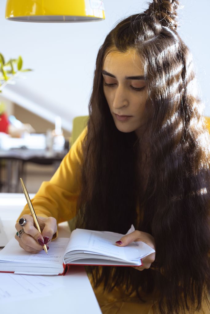 A young woman with long hair sits at a desk, writing in a notebook with a gold pen. She wears a yellow shirt and is focused on her writing.