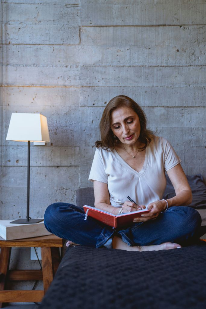 A woman sitting cross-legged on a bed, writing in a red notebook. She has medium-length brown hair and is wearing a light-colored shirt. There is a lamp on a wooden table beside her, and the background features a textured concrete wall.