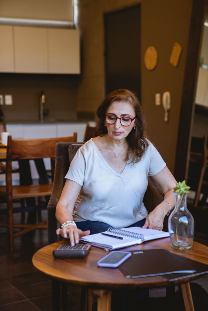 A woman sitting at a wooden table in a cafe, using a calculator and reviewing notes in a notebook, with a laptop and a potted plant on the table.
