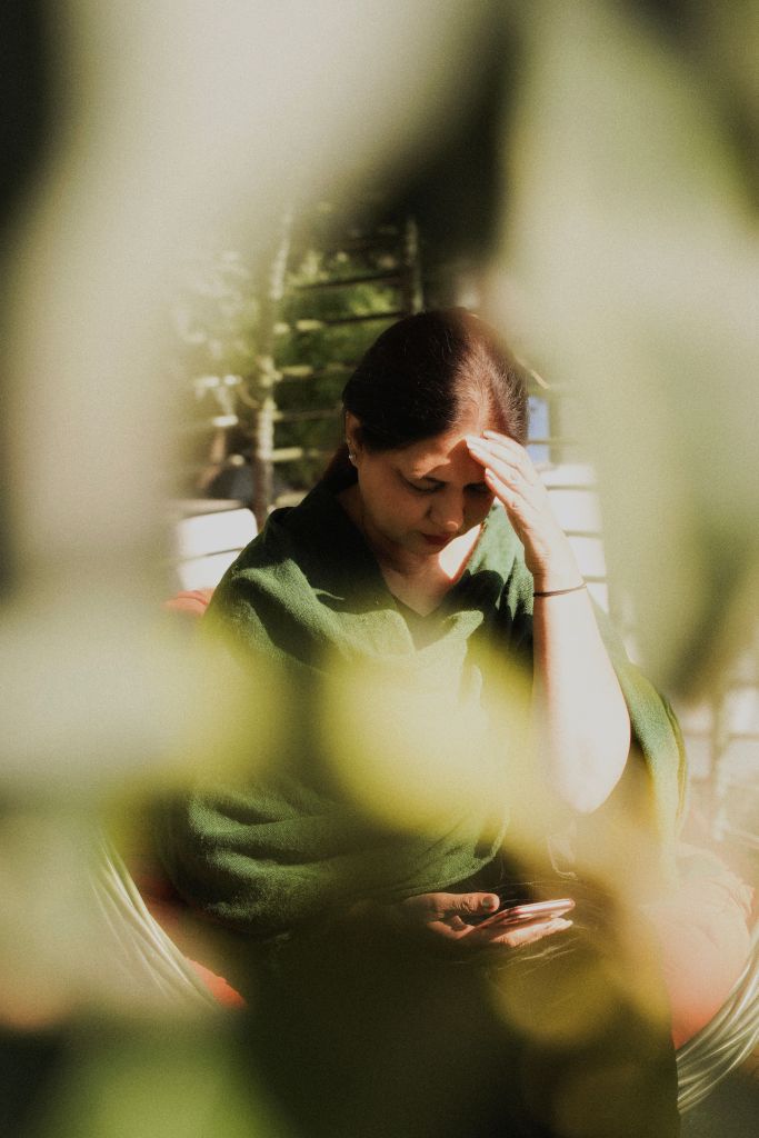 A woman sitting outdoors, wearing a green shawl, with a pensive expression while looking at her phone, partially obscured by leaves.