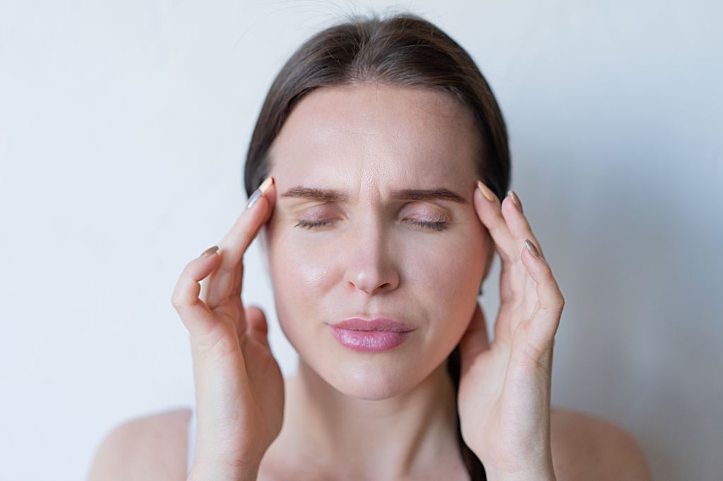 A woman with a neutral expression is holding her temples with both hands, eyes closed, appearing to be in discomfort or experiencing a headache, against a light background.