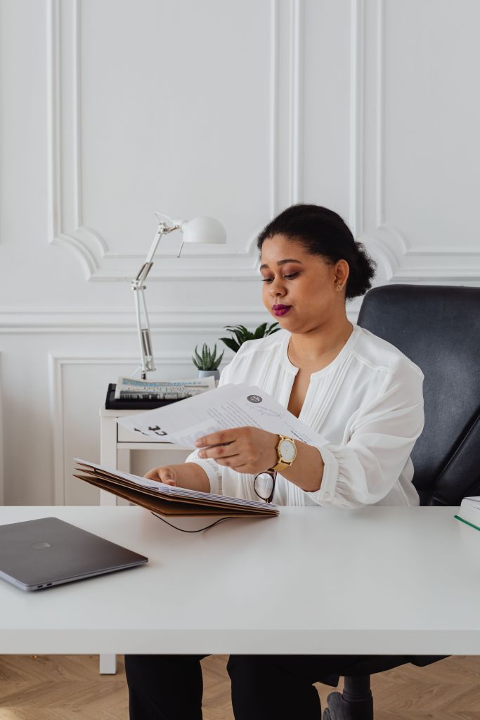 A woman sitting at a desk, reviewing documents while wearing a white blouse and glasses, with a laptop and a plant in the background.