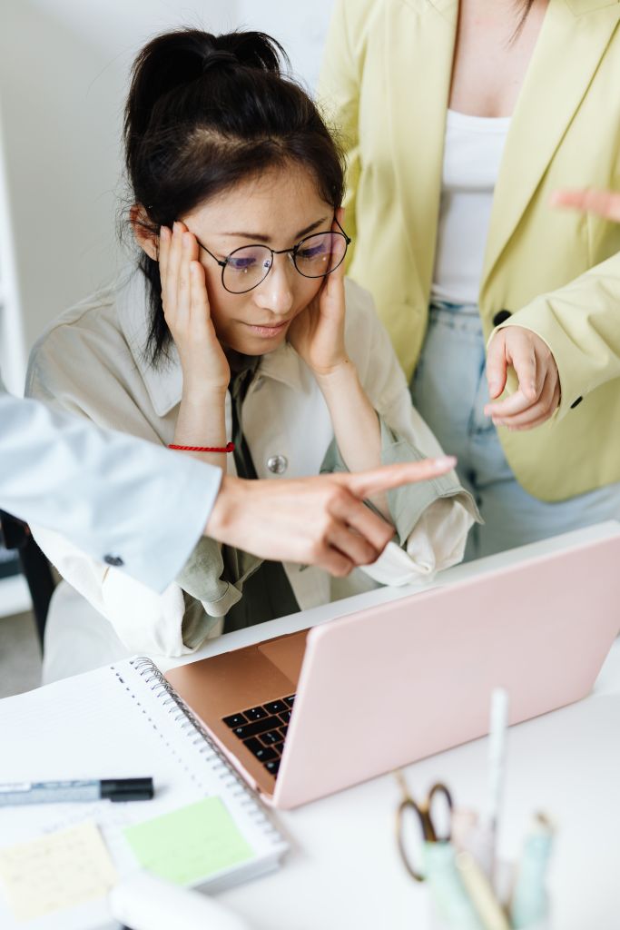 A young woman with glasses looks stressed while working on a laptop, with her hands on her face. Two other individuals in the background are pointing and engaged in conversation.