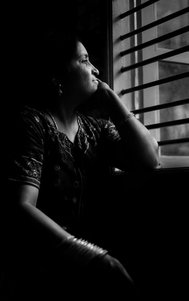 A woman resting her chin on her hand, gazing thoughtfully out of a window with slatted blinds, captured in black and white.