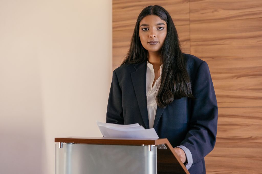 A woman in a black blazer stands behind a podium, holding papers and looking confidently at the camera.