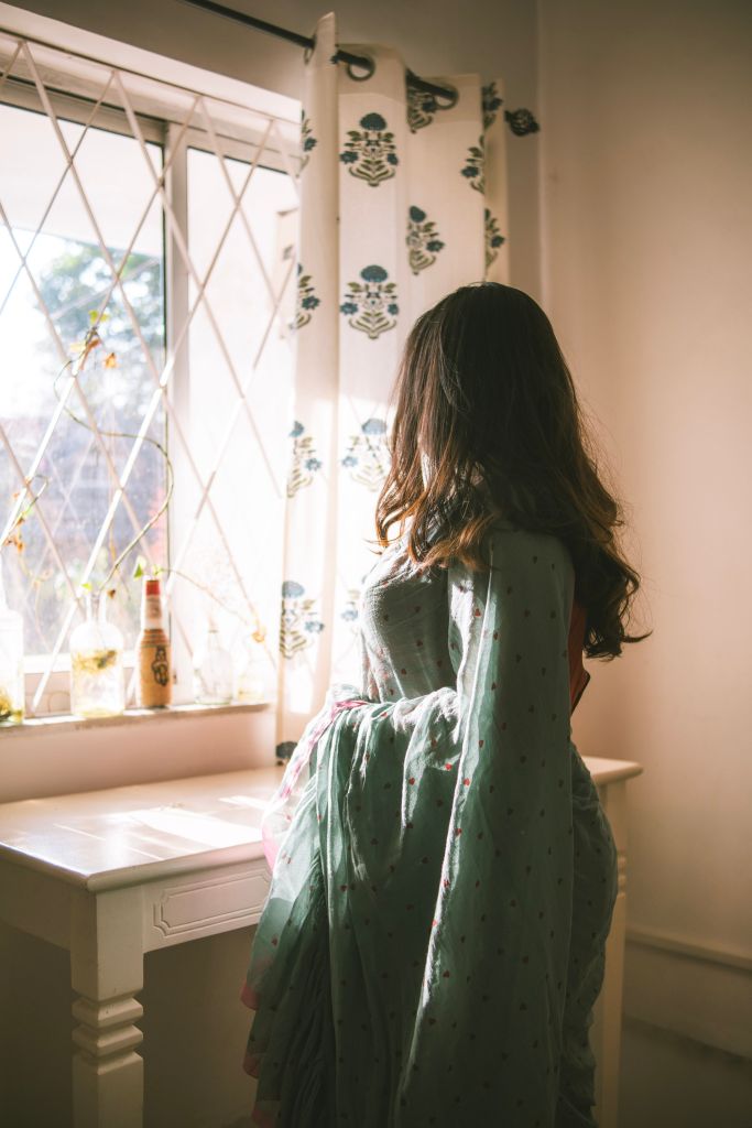A woman standing by a window, wearing a green saree with floral patterns, looking out. Sunlight is streaming in, illuminating the room and a white table with decorative items.