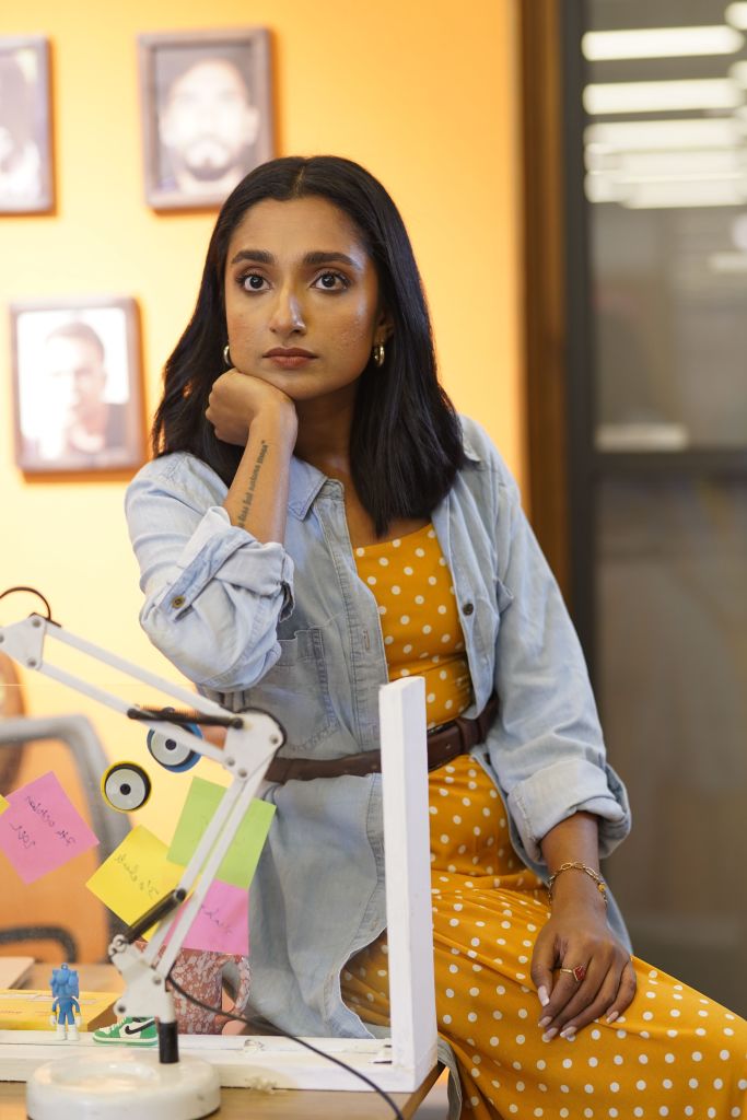 A young woman sitting at a desk with colorful sticky notes, wearing a denim jacket over a yellow polka dot dress, looking thoughtfully at the camera.