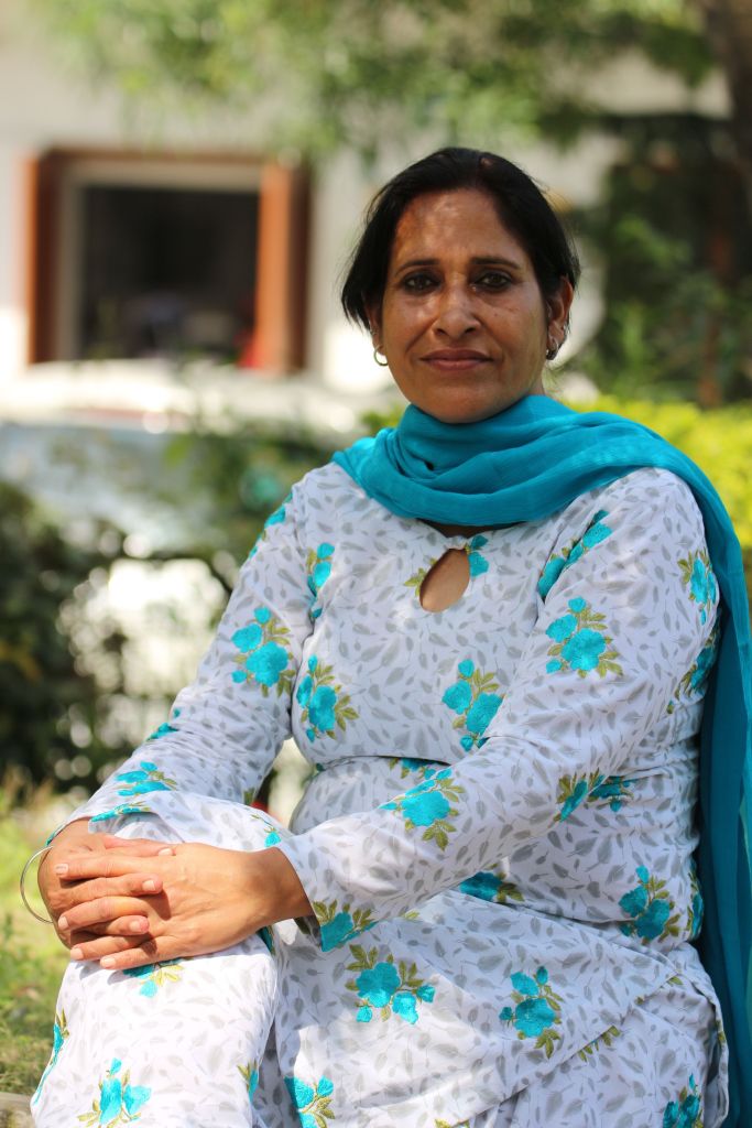 A woman dressed in a floral-patterned kurta and a blue scarf sitting outdoors with a serene expression.