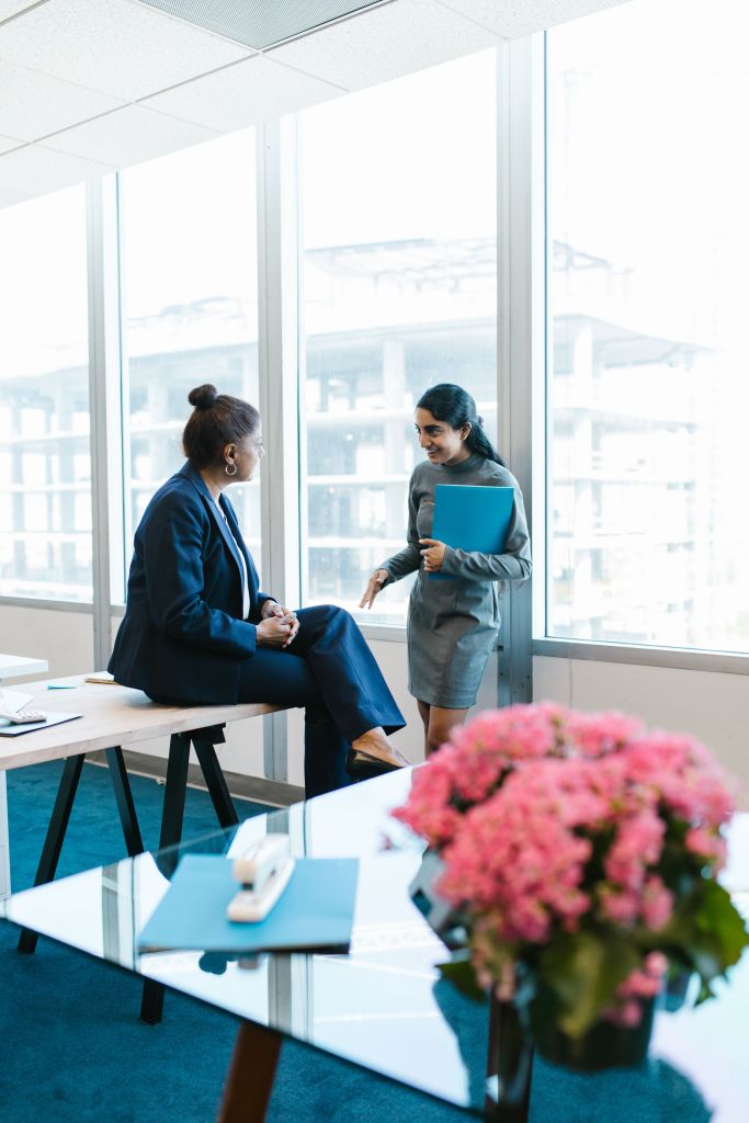 Two businesswomen engaging in conversation in an office setting, one sitting on a bench and the other standing holding a folder, with large windows showing a construction site in the background.