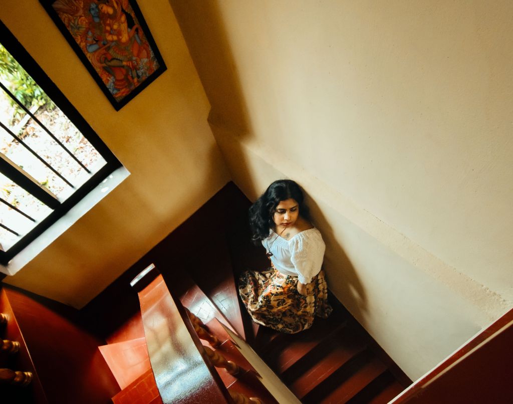 A woman wearing a white blouse and patterned skirt sitting on stairs, with a view of a window and artwork on the wall.