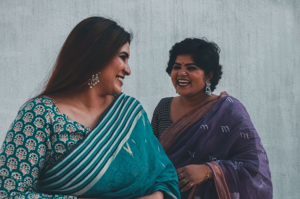 Two women wearing traditional attire smile and chat against a neutral background.