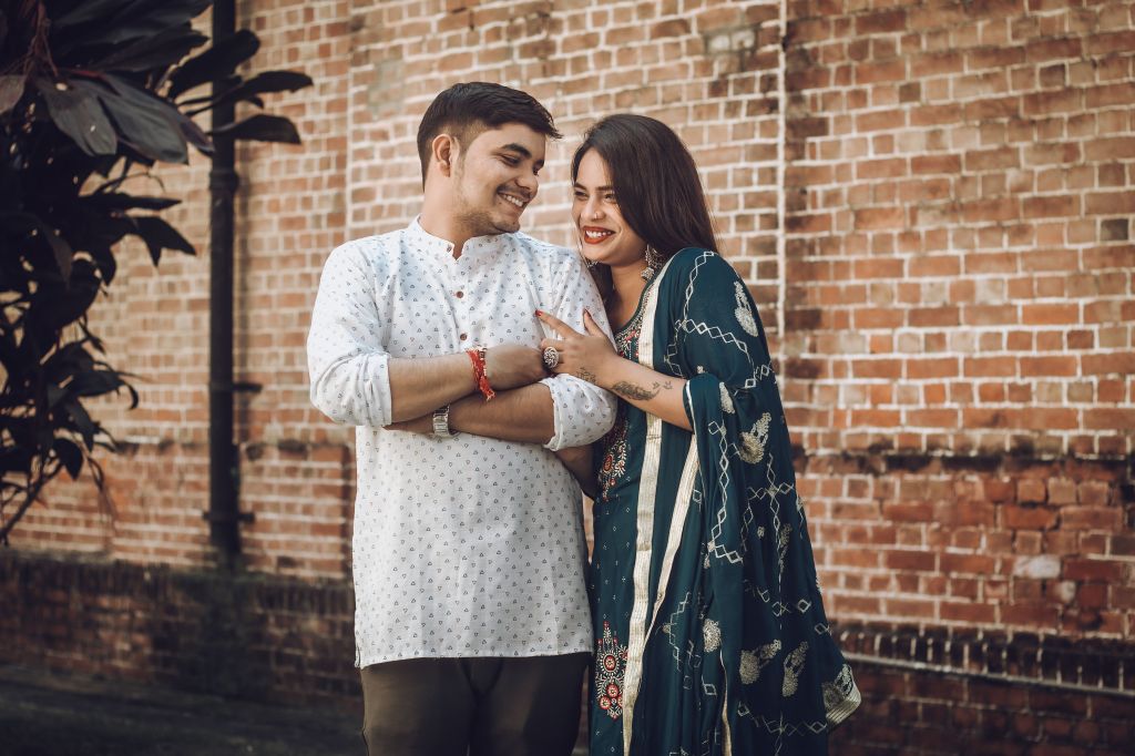 A smiling couple posing together outdoors against a brick wall, dressed in traditional attire. The man wears a white patterned kurta, while the woman is in a teal outfit with intricate designs.