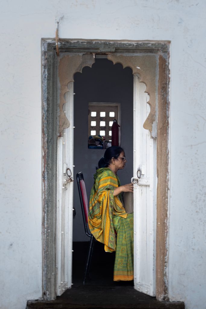 A woman in a green traditional attire is sitting inside a doorway, partially visible. The doorway has an ornate arch design, revealing a glimpse of a room with a window and some items in the background.