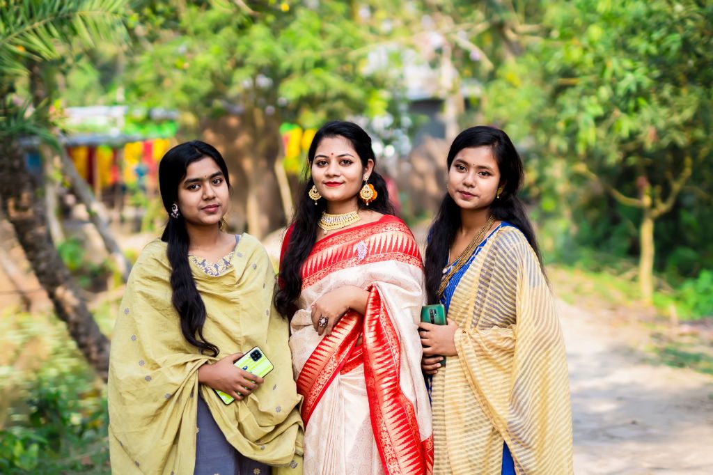 Three women standing together outdoors, wearing traditional attire, with a background of greenery and colorful decorations.