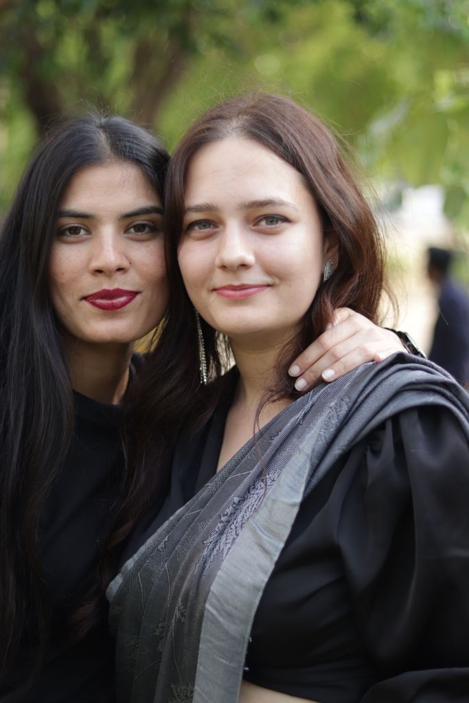 Two women posing closely together with friendly smiles, one wearing a black outfit and the other draped in a grey saree, against a green outdoor background.