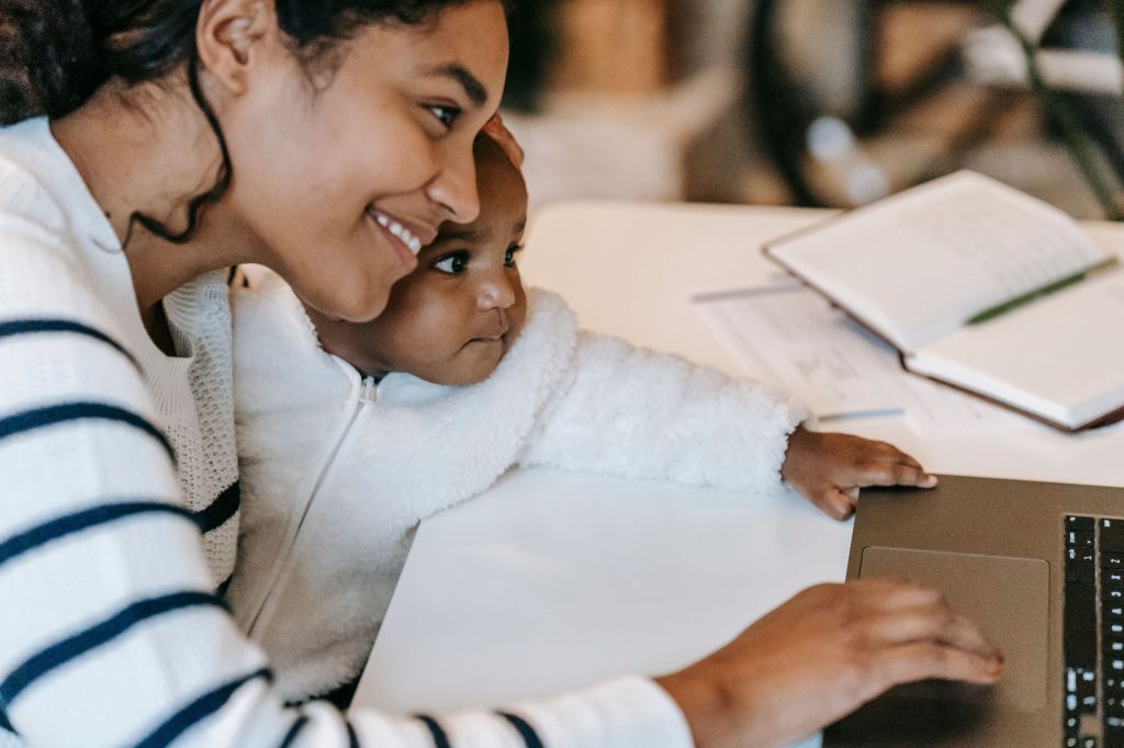 A smiling woman sitting at a table, using a laptop, while a baby beside her curiously looks at the screen.