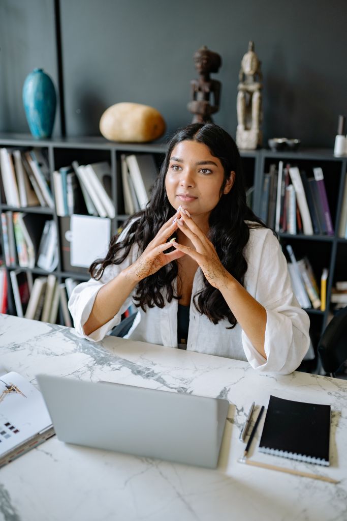 A woman with long, dark hair sits at a marble desk, looking thoughtfully at a laptop. She has her hands clasped together and various decorative items and books can be seen in the background.