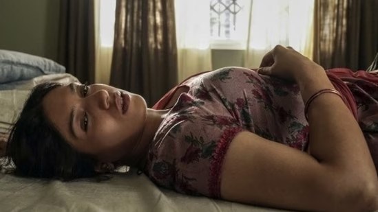 A woman lies on a bed, looking contemplative with a slightly worried expression, surrounded by soft natural light from a nearby window.
