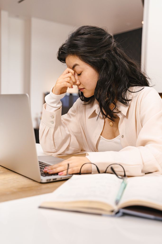 A woman with dark, wavy hair sits in front of a laptop, looking stressed while rubbing her forehead. Notebooks and a pen are visible on the table.