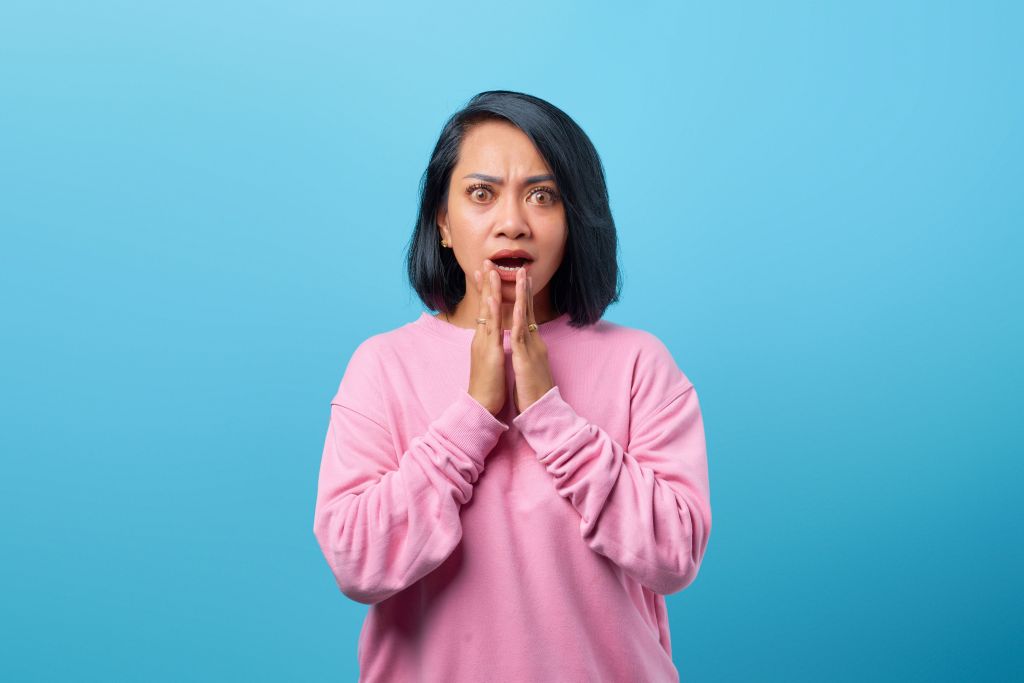 A woman in a pink sweatshirt looks surprised and shocked, with her hands clasped together in front of her mouth, against a blue background.