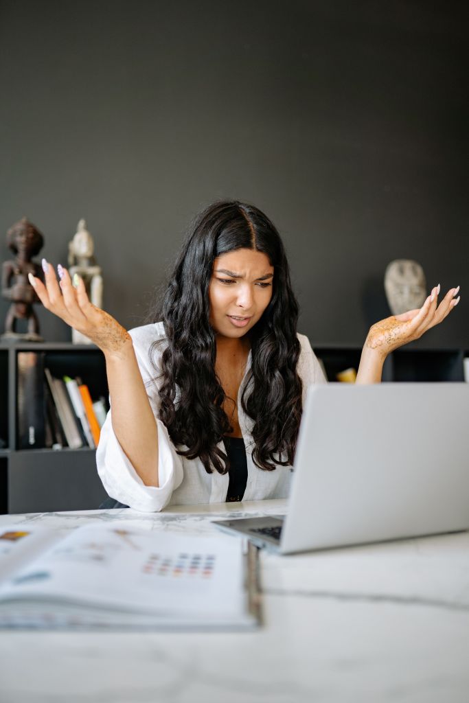 A woman with long, wavy hair is sitting at a desk in front of a laptop, looking frustrated and throwing her hands up in exasperation.